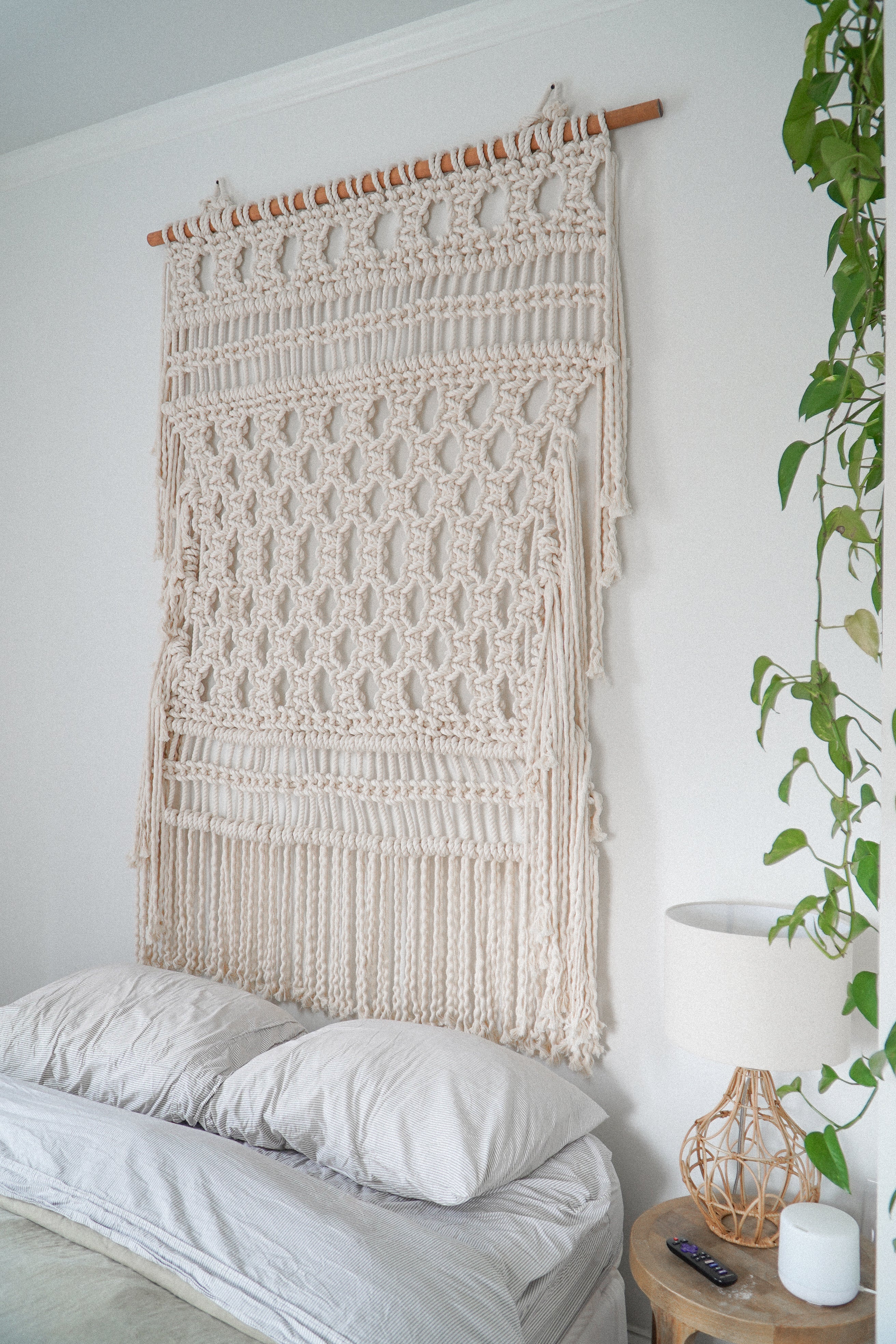 A large cream macrame wall hanging suspended from a wooden rod above a bed in a bright bedroom. A white nightstand with a lamp and plant is visible on the right.